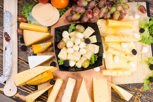 Photo gourmet display of different cheeses arranged around a wooden board on a buffet table with a central bowl filled with diced cheese, cocktail onions and olives, overhead view