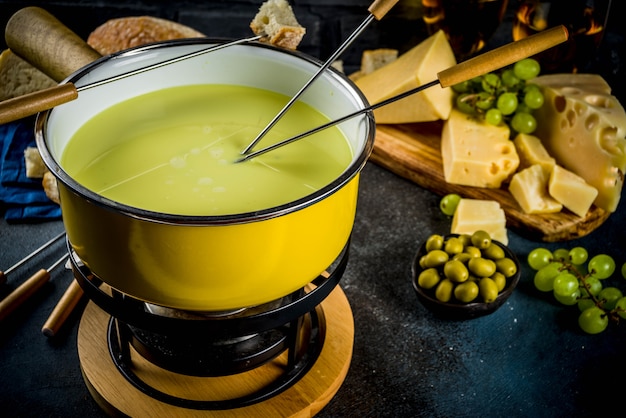 Gourmet Swiss fondue in traditional fondue pot, with forks, various cheeses, olives, bread and grape