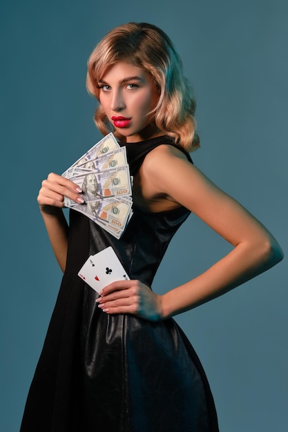 Graceful blonde lady with bright makeup in black stylish dress is holding some money and cards while posing sideways against blue studio background Gambling entertainment poker casino Closeup