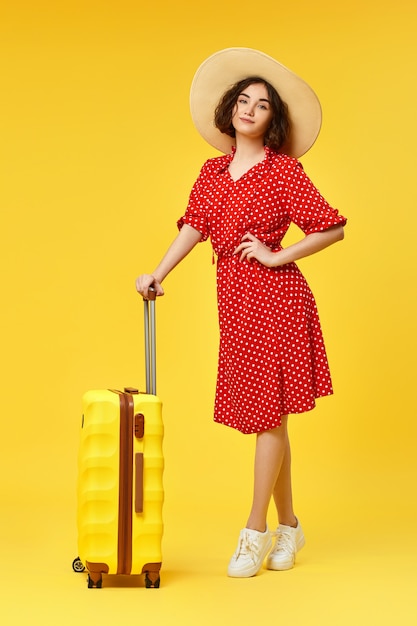 Graceful curly woman in red dress and hat with suitcase going traveling on yellow background.