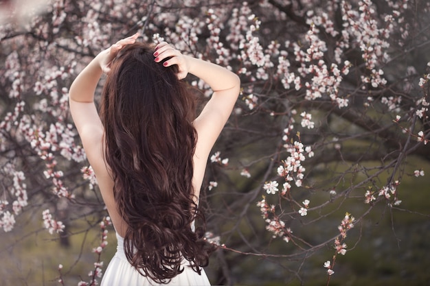 Graceful female in white dress standing with her back under flowering trees in spring garden