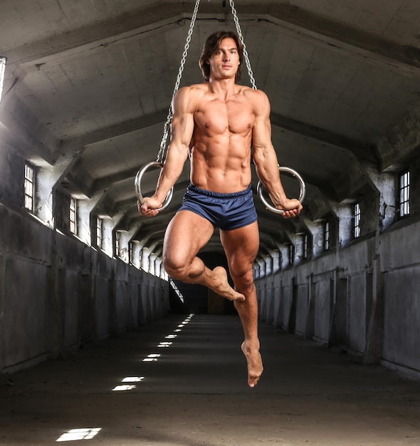 Graceful muscular sportsman workouts in the air with gymnastic\
rings, training in an abandoned industrial building