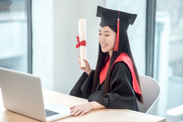졸업장으로 졸업하십시오. 그녀의 졸업장을 들고 화상 통화를하는 귀여운 아시아 웃는 대학원