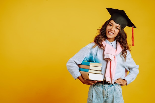 Graduate woman in a graduation hat on her head, with books on yellow.