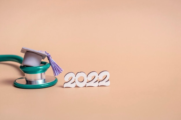 Graduation cap with stethoscope and numbers 2022 on beige background, close up with copy space. Medical education concept.