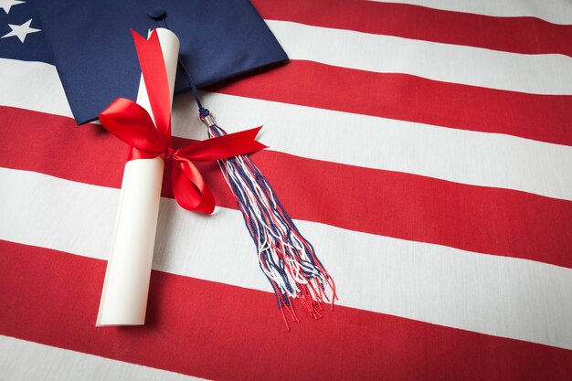 Foto cappello da laurea con nappa e diploma avvolto in nastro rosso appoggiato sulla bandiera americana