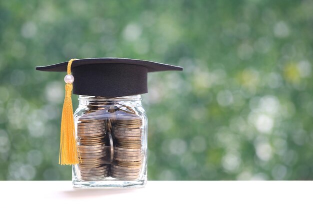 Graduation hat on coins money in the glass bottle on natural green background Saving money for education concept