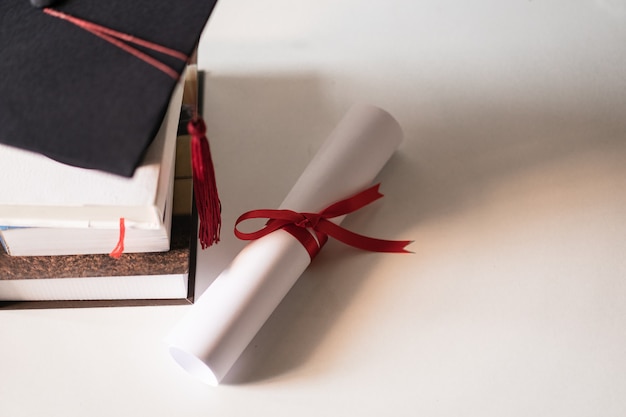 A graduation hat or mortarboard and diploma certificate paper\
tied with red ribbon on a stack of books with empty space slightly\
undersaturated with vignette for vintage effect