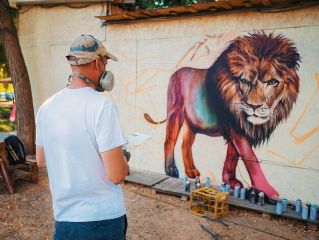 写真 落書きアーティストが通りの壁に絵を描くエアゾール スプレー ボトルを持つ男カラフルなライオンを描く保護マスクの若い才能のある男