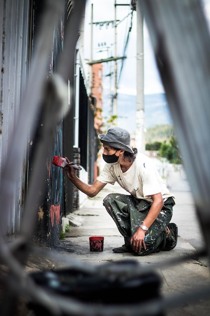 Foto graffitikunstenaar met een gezichtsmasker en met verf besmeurde kleding schildert met een penseel op een straatmuur