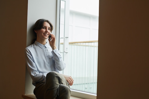 Grafisch portret van lachende jonge zakenvrouw spreken door smartphone zittend op de vensterbank op het werk, kopieer ruimte