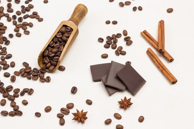 Grains of coffee in wooden scoop and on table. Chocolate, cinnamon sticks and star anise. White background. Top view