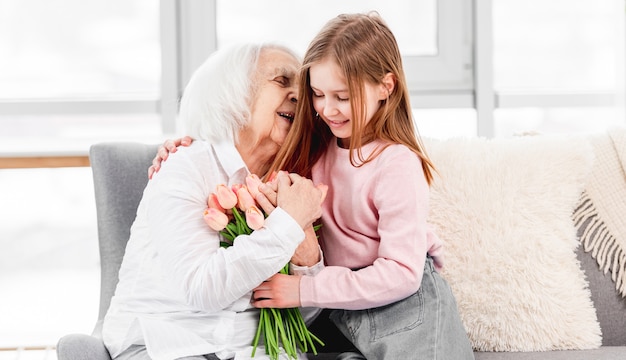 Grandaughter gives flowers tulips bouquet to grandmother and hug her indoor at home