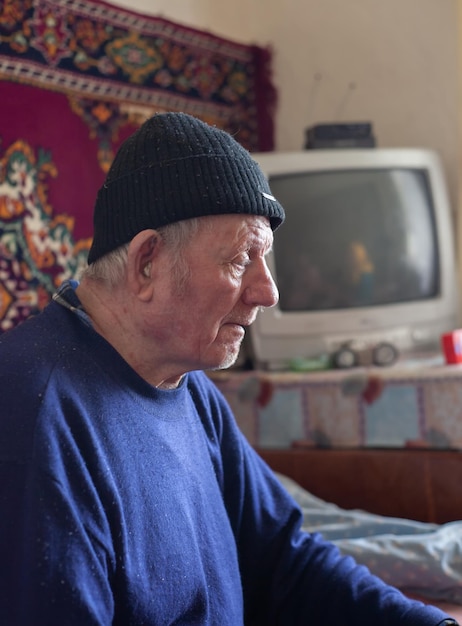 Grandfather an elderly man sits in a house in a cap Blurred background of old carpet and TV