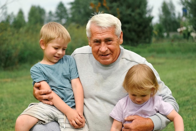 Grandfather and grandchildren have fun during walk In Park Happy family time Old man grandpa hugging children boy and girl Summer day Smiling Senior male spending time with his grandkids together