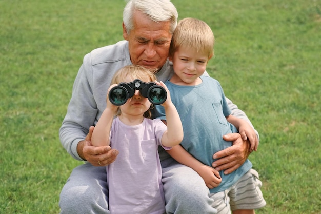 Grandfather and grandchildren using binoculars In Park Happy family time Old man grandpa playing with children boy and girl Summer day Smiling Senior male spending time with his grandkids together