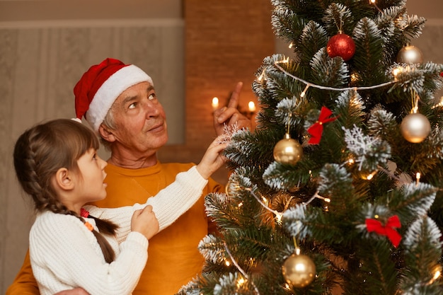 Nonno e nipote che decorano l'albero di natale, famiglia che indossa abiti in stile casual guardando il bellissimo albero di z mas, festeggiando insieme il capodanno.