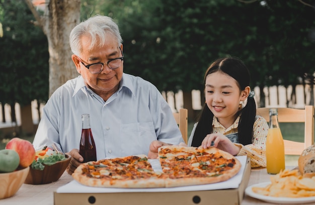 家の庭でピザを食べている祖父と孫娘。夏休みの家族との定年ライフスタイル。