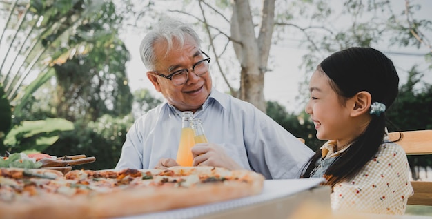 Дед и внучка с пиццей в саду дома. Образ жизни пенсионного возраста с семьей на летних каникулах.