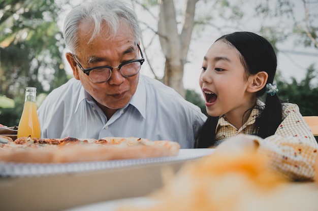 Дед и внучка с пиццей в саду дома. Образ жизни пенсионного возраста с семьей на летних каникулах.