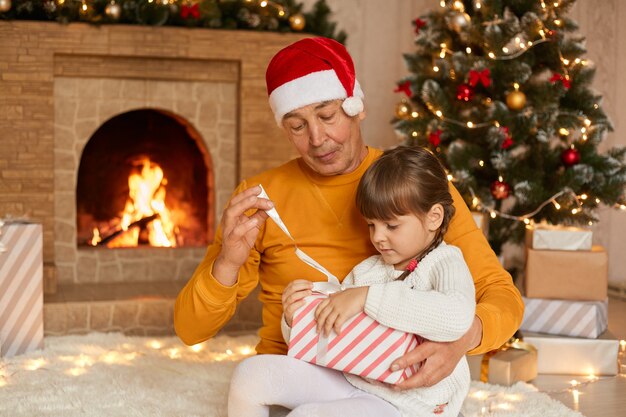 Grandfather and granddaughter opening Christmas presents, posing in living room with new year decoration, kid sitting on man's knees, being concentrated, look at box, posing near fireplace.