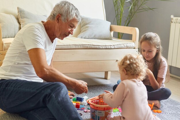 リビングルームのソファの近くの床に座っておもちゃで遊ぶ祖父と孫娘が、家の内部でゲームを楽しんで一緒に幸せな時間を過ごします