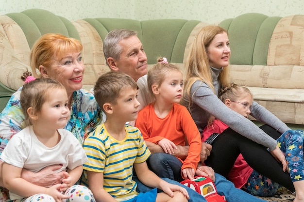 Дедушка и бабушка с дочерью и внуками смотрят телевизор, сидя на полу у дивана