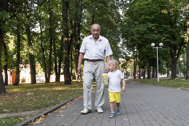 Grandfather and grandson are walking in the park along the path with a glass of popcorn An old man and a little boy have snacks on the go