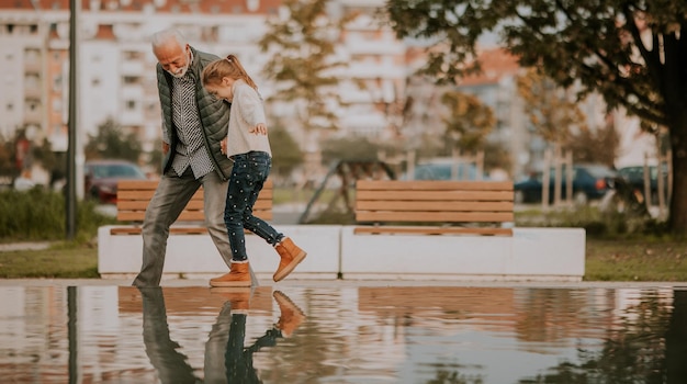 Grandfather spending time with his granddaughter by small water pool in park on autumn day