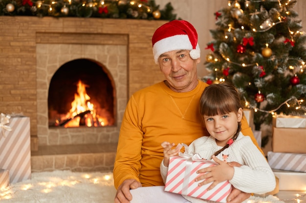 Grandfather with her granddaughter sitting in living room and , child holding present box, posing on floor on soft carpet near fir tree and fireplace.