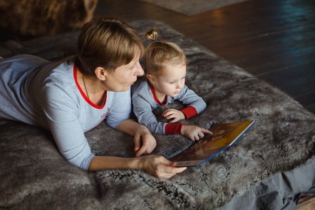 Photo grandma and granddaughter have fun together reading a book on the bed .