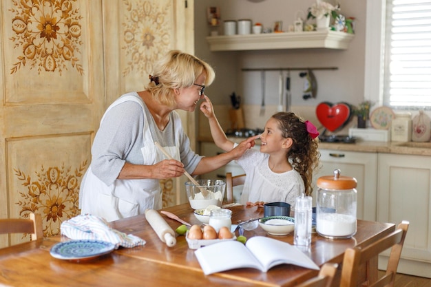 Grandma in white apron with cute curly granddaughter spend wonderful time in the kitchen, smiling and there are happy. Horizontal view.