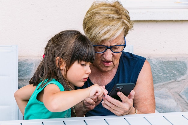 사진 할머니와 손녀가 테라스에서 휴대폰을 보고 놀고 있습니다. 가족, 손자, 조부모 및 기술 개념.