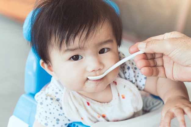 Grandmother feeding cute asian baby girl with a spoon at home. 