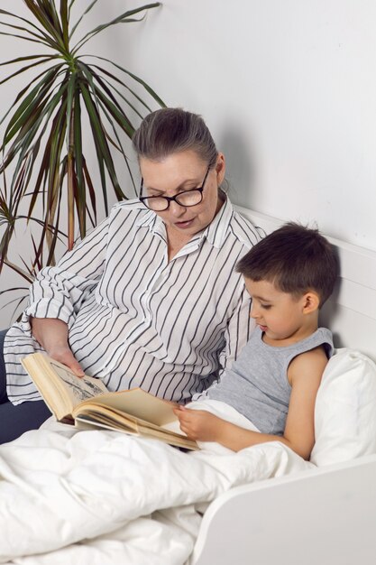 Photo grandmother in glasses and a white shirt reads a book to her grandson lying on the bed in a white children's room