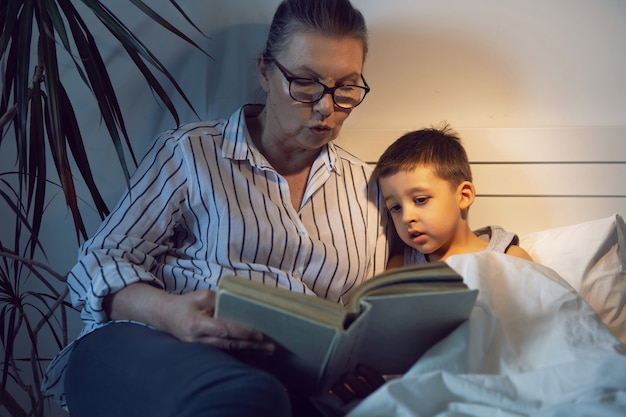 Grandmother in glasses and a white shirt reads a book to her grandson lying on the bed in a white children's room