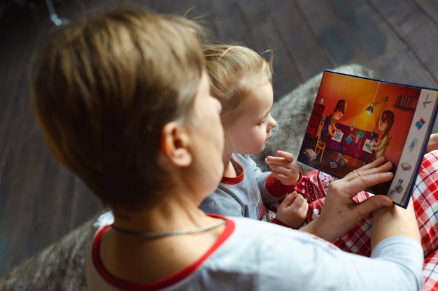 Grandmother and granddaughter sit on bed in pajamas and reading book