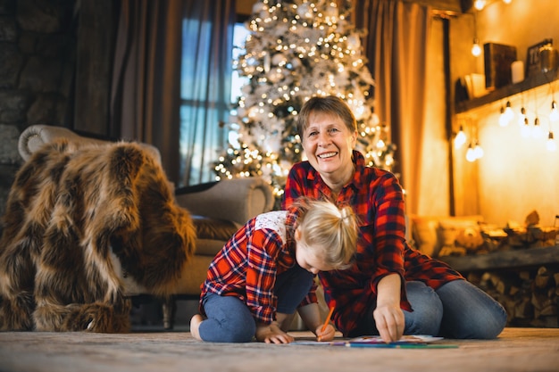 Photo grandmother and granddaughter sitting on the carpet in front of the tree and draw.