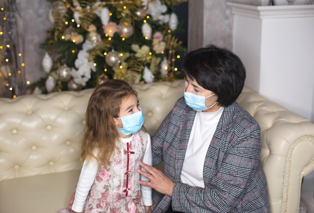 Grandmother and granddaughter on the sofa in the living room with Christmas decor hugging in medical masks on their faces. A family holiday during the outbreak of coronavirus and disease. New Year