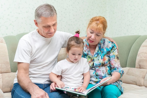 Grandmother and grandfather are reading a book to their granddaughter while sitting on the couch