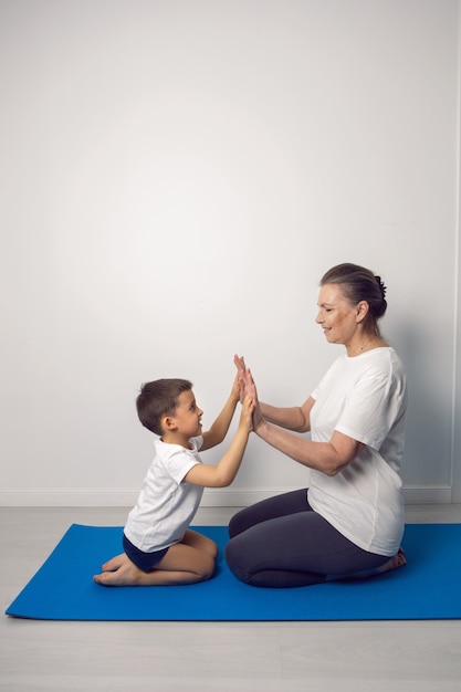 Grandmother and her grandson are sitting on a yoga mat in a white apartment