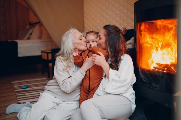 Grandmother Mother and child kissing and playing on sofa near fireplace Mom and baby Parent with daughter and grandson little kid relaxing at home Family having fun together Mother's day