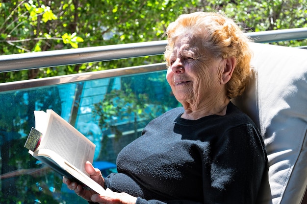 Grandmother sitting on the balcony of house reading a book.