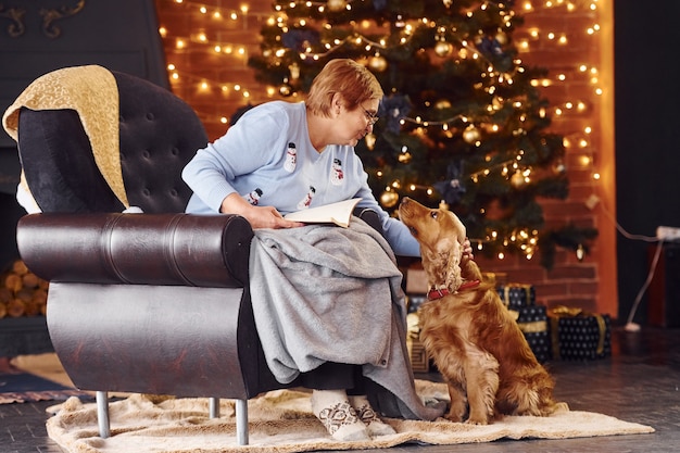 Grandmother with book in hand indoors with dog in christmas decorated room.