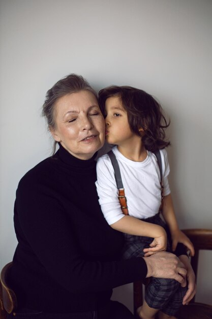 Foto la nonna con un nipote di quattro anni con i capelli lunghi si trova contro un muro bianco con una sedia