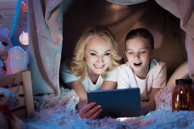 Grandmother with girl are watching video in blanket house at night