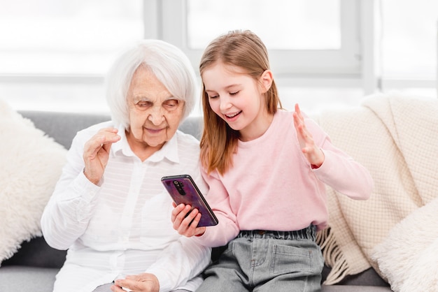 Nonna con la nipote seduta sul divano e parlando online utilizzando lo smartphone