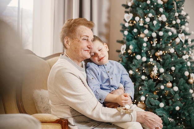 Grandmother with her grandson on Christmas day. Old woman and little boy decorating Christmas tree. Woman and boy wearing beige and blue clothes.