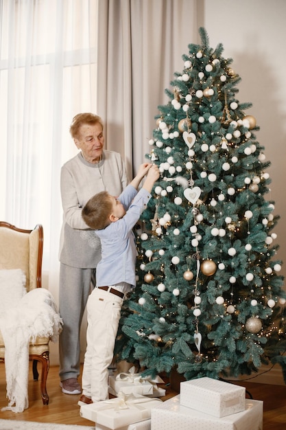 Grandmother with her grandson on Christmas day. Old woman and little boy standing and decorating Christmas tree. Woman and boy wearing beige and blue clothes.