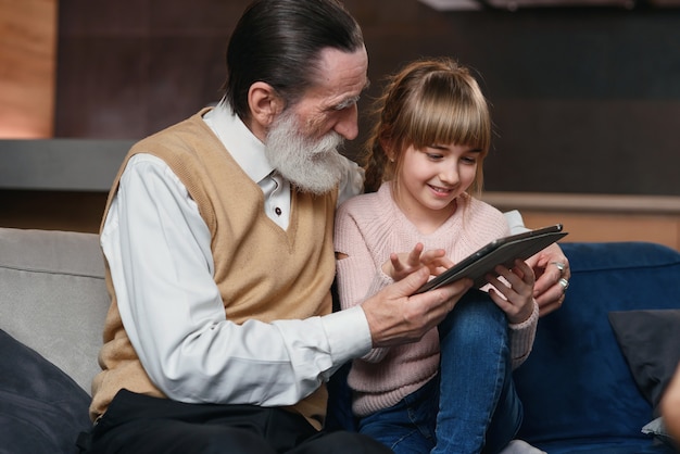 아늑한 집에서 태블릿 pc를 사용하여 그의 손녀와 할아버지. 어린 소녀는 할아버지에게 스마트 기기를 사용하도록 가르치십시오.